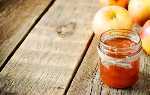 Zu sehen ist ein Holztisch, auf dem ein paar Äpfel liegen und ein Glas mit Apfelmarmelade steht.