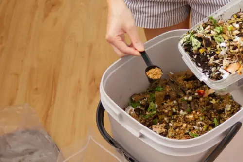 Zu sehen ist eine Peron, die Küchenabfälle in einen Bokashi-Eimer gibt.