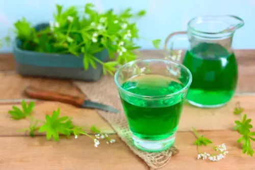 Auf diesem Bild steht eine kleine Kiste mit frischem Waldmeister neben einer Karaffe mit Waldmeistersirup im Hintergrund auf einem Holztisch. Vorne ist ein Glas Waldmeisterlimonade zu sehen. [Foto: AdobeStock_rainbow33]