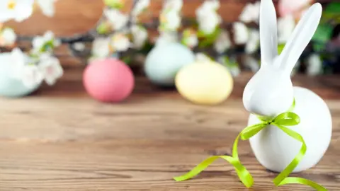 Eine Hasenfigur aus weißem Porzellan mit einer grünen Schleife um den Hals steht auf einem Holztisch, im Hintergrund bunte Ostereier und Blüten
