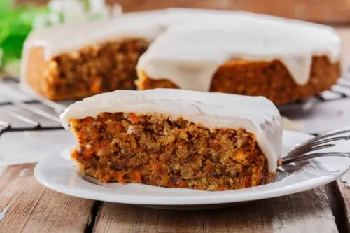 Rueblikuchen mit Frischkaese-Creme auf einem Teller. Im Hintergrund ist der ganze Kuchen zu sehen, waehrend ein einzelnes Stueck im Vordergrund erkennbar ist. [Foto: AdobeStock_koss13]