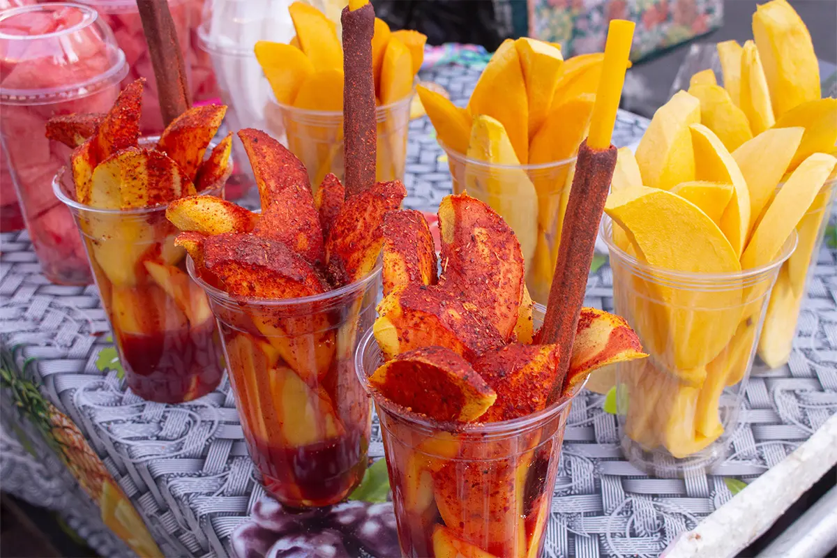 Frutas Locas auf einem Tisch: Mango- und Wassermelonenstücke mit Chamoy-Sauce und Gewürzen in Bechern. In der vorderen Reihe stehen die servierfertigen Becher, dahinter stehen Becher mit Früchten ohne Gewürz und Sauce. Darunter ist eine schwarz-weisse Tischdecke zu erkennen. [Foto: AdobeStock_DAVID]