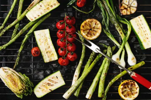 Diverse Grillgemüse (hier Spargel, Zucchini, Rispentomaten, Lauch) liegen auf einer dunkeln Grillplatte nebeneinander. [Foto: AdobeStock_Natasha Breen]