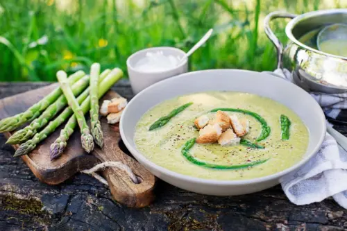 Spargelcremesuppe, die auf einem Gartentisch in einem weissen Teller angerichtet wurde. In der Suppe befinden sich gekochte Spargelstangen und geröstetes Brot. Daneben liegen grüne Spargelstangen, Brot und es ist ein Gefäß mit Crème fraîche zu sehen. [Foto: AdobeStock_Dušan Zidar]