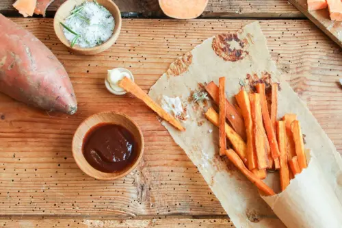 Suesskartoffel-Pommes liegen auf Backpapier neben Dips, Salz und Pfeffer auf einem rustikalen Holztisch. [Foto: AdobeStock_eflstudioart]