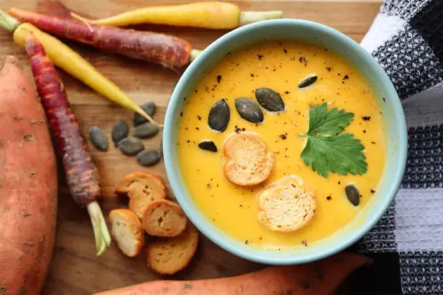 Ansicht von oben auf eine hellblaue Schale mit cremiger Süßkartoffelsuppe, die von einer rohen Süßkartoffel sowie bunten Möhren umgeben ist. Im Hintergrund eine Holzoberfläche und ein schwarzweiß kariertes Küchentuch. [Foto: AdobeStock_Adrian]