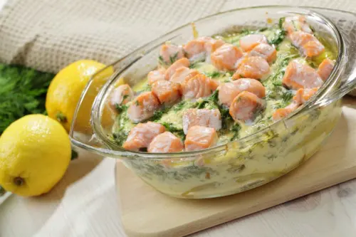 Lachs aus dem Ofen in einer Auflaufform aus Glas – hier serviert auf einer cremigen Dill-Senf-Sauce und Kartoffeln. Neben der Auflaufform, die auf einem Holzbrettchen steht, liegen frische Zitronen. [Foto: AdobeStock_StockphotoVideo]