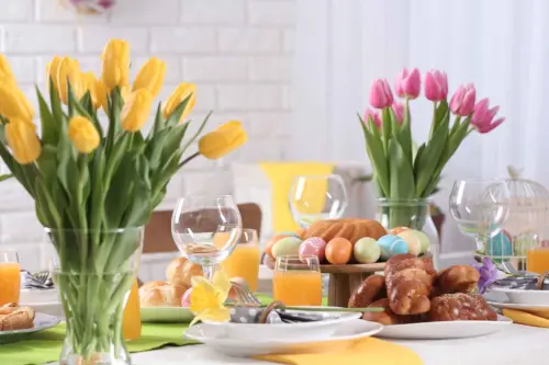 Festliche Tafel gedeckt mit bunten Oster-Rezepte wie bemalten Eiern in einem Korb, Hefegebaeck, Kuchen und orangefarbenem Fruchtsaft. Dazwischen gehen Vasen mit Gelben und Pinken Tulpen. [Foto: AdobeStock_New Africa]