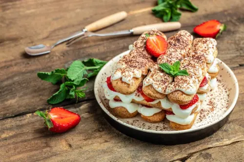Erdbeer-Tiramisu aus einem weissen Teller, der auf einem rustikalen Holztisch steht. Daneben liegen eine Erdbeere sowie Holzbesteck. Das Tiramisu aus Löffelbisquit, Erdbeeren und Mascarpone ist bestreut mit Kakaopulver. [Foto: AdobeStock_Надія Коваль]