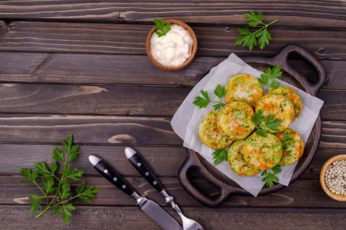 Gemüsefrikadellen liegen auf einer rustikalen Servier-Platte. Diese steht auf einem Holztisch. Daneben sind Messer und Gabel, etwas frische Petersilie und ein kleines Schälchen mit Mayonnaise zu sehen. [Foto: AdobeStock_Veronika Idiyat]