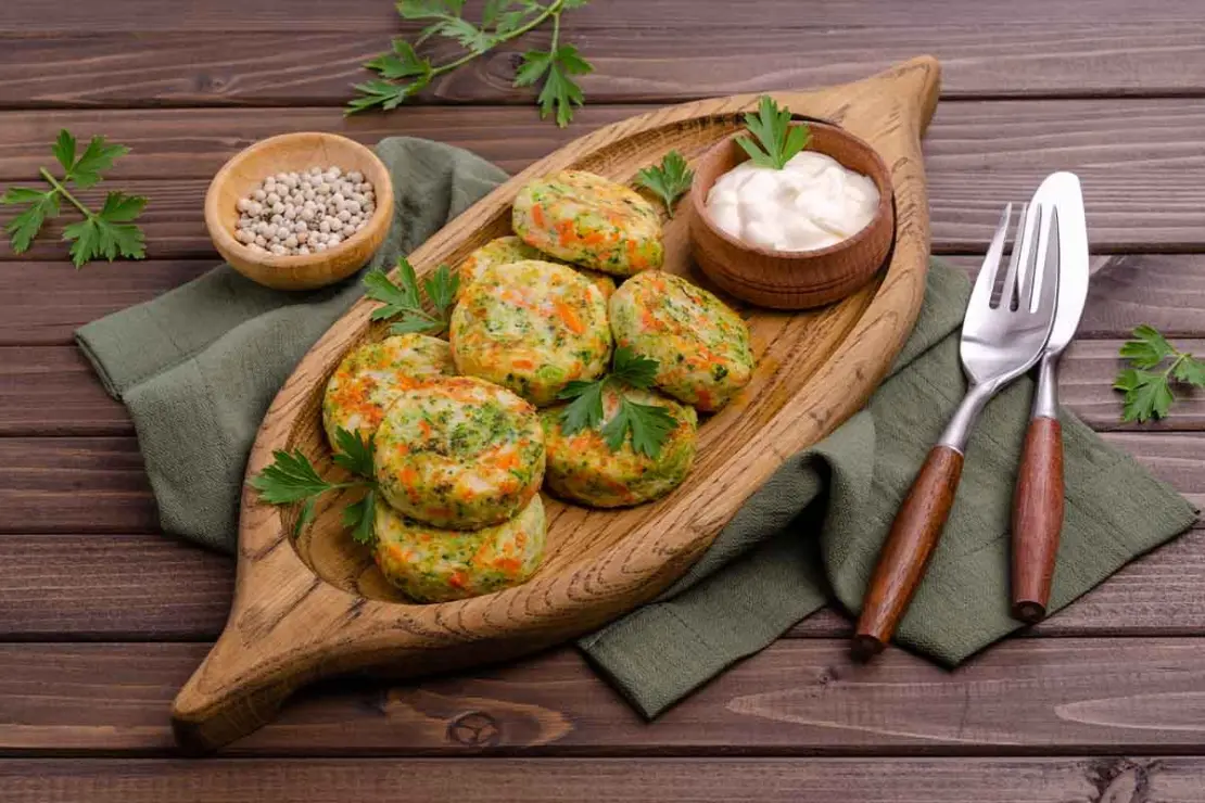 Gemüsefrikadellen liegen auf einem Servier-Tablett aus Holz, auf dem auch ein kleines Schälchen mit Mayonnaise steht. Neben dem Tablett liegen ein Messer, eine Gabel und etwas grober weißer Pfeffer in einem kleinen Schälchen. Das Tablett steht auf einer dunkelgrünen Leinenserviette auf einem Holztisch. [Foto: AdobeStock_Veronika Idiyat]