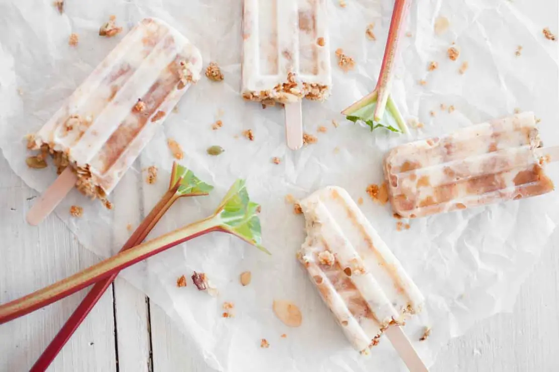Joghurt-Eis mit Rhabarberstueckchen, das am Stiel gefroren wurde, liegt nebeneinander auf einem weissen Backpapier neben Rhabarber-Stangen auf einem weissen Holztisch. Das Eis ist eine gute Moeglichkeit, eine erfrischende Variante des Stielgemueses zu kreieren, wenn Sie Rhabarber zubereiten moechten. [Foto: AdobeStock_Kati Finell]