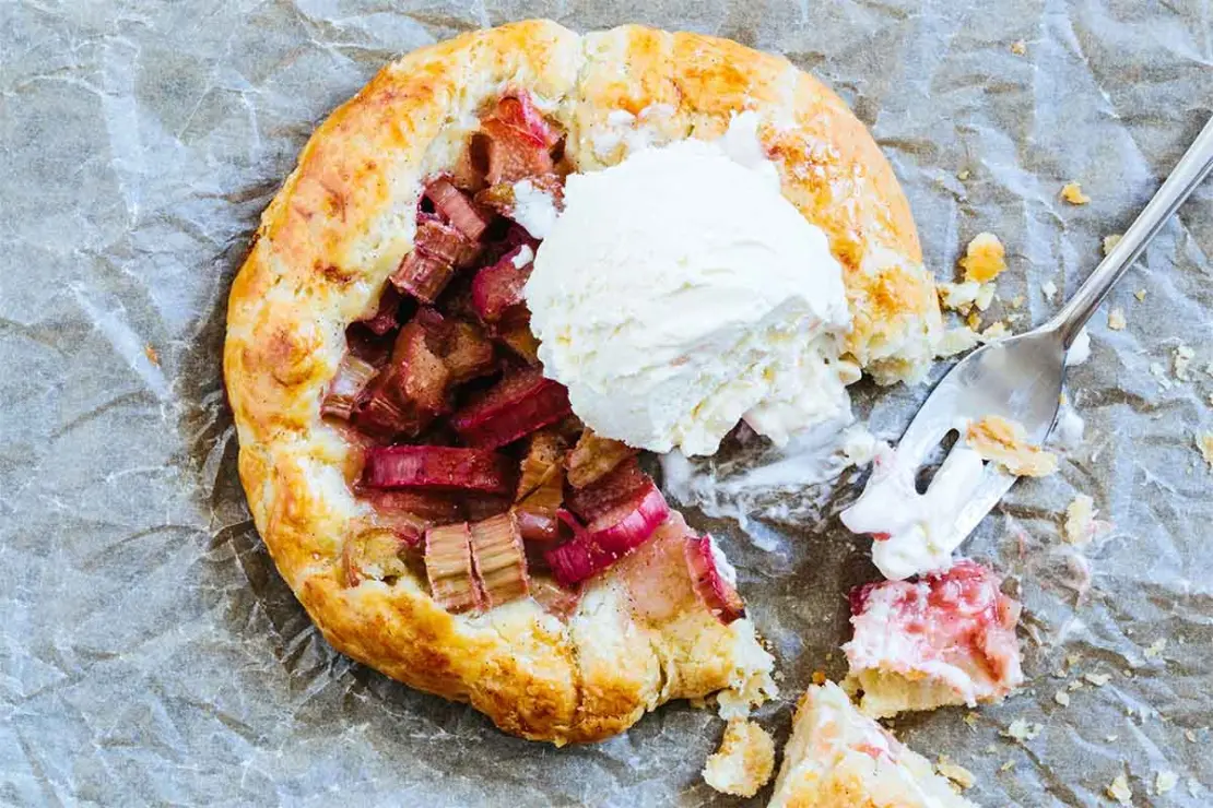 Eine knusprige Galette mit Rhabarberfuellung liegt auf etwas Backpapier auf einem Tisch. Oben auf der mit Rhabarber befuellten Mitte liegt eine Kugel mit leckerem Vanilleeis. [Foto: AdobeStock_Peti]
