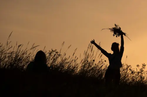 Frau mit einem gebundenen Strauß in der Hand tanzt auf einem Feld im Gegenlicht der untergehenden Sonne. Links von ihr sitzt eine weitere Person im Feld.