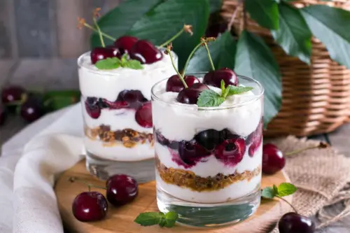 Geschichtetes Kirsch-Dessert in Gläsern. Zu sehen sind verschiedene Schichten aus Joghurt-Creme, Kirschen und einer Knuspermischung. Auf der oberen Schicht zieren Kirschen und Minzblätter die Süßspeise. [Foto: AdobeStock_qwartm]
