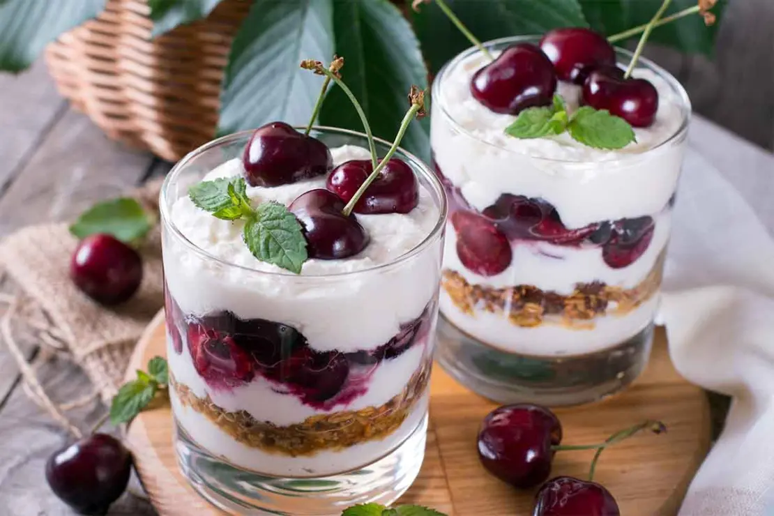 Zwei geschichtete Kirsch-Desserts stehen nebeneinander auf einem Holzbrettchen. Oben auf den Schichten aus Joghurt-Creme, Kirschen und Knusperbröseln tronen liebevoll etwas Minze und jeweils drei Kirschen zur Zierde. [Foto: AdobeStock_qwartm]