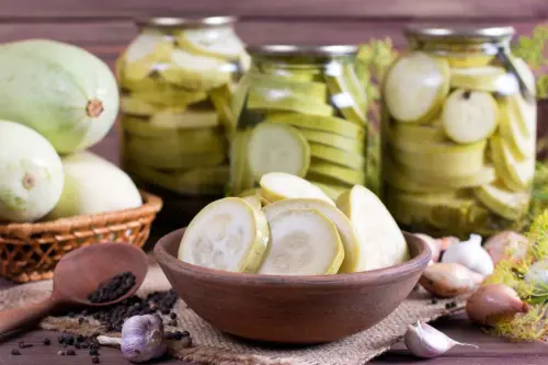 Eingelegte Zucchinischeiben in einer Schüssel vor Einmachgläsern mit Zucchinisalat. Daneben liegen frische Zucchini, Knoblauch, Pfefferkörner und Schalotten auf einem rustikalen Holztisch. [Foto: AdobeStock_qwartm]