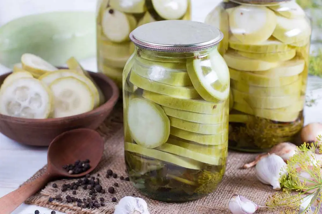 Mehrere Gläser mit eingelegten Zucchinischeiben stehen auf einem Tisch. Im Vordergrund liegt ein Holzlöffel mit Pfefferkörnern, daneben frischer Knoblauch, Dill und Schalotten. [Foto: AdobeStock_qwartm]
