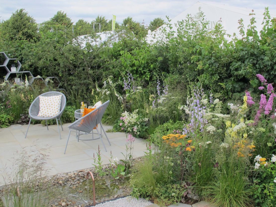 Ein befestigter Sitzplatz im Garten, von bienenfreundlichen Pflanzen umgeben und mit einem flachen, mit Kieseln ausgelegten Wasserlauf.