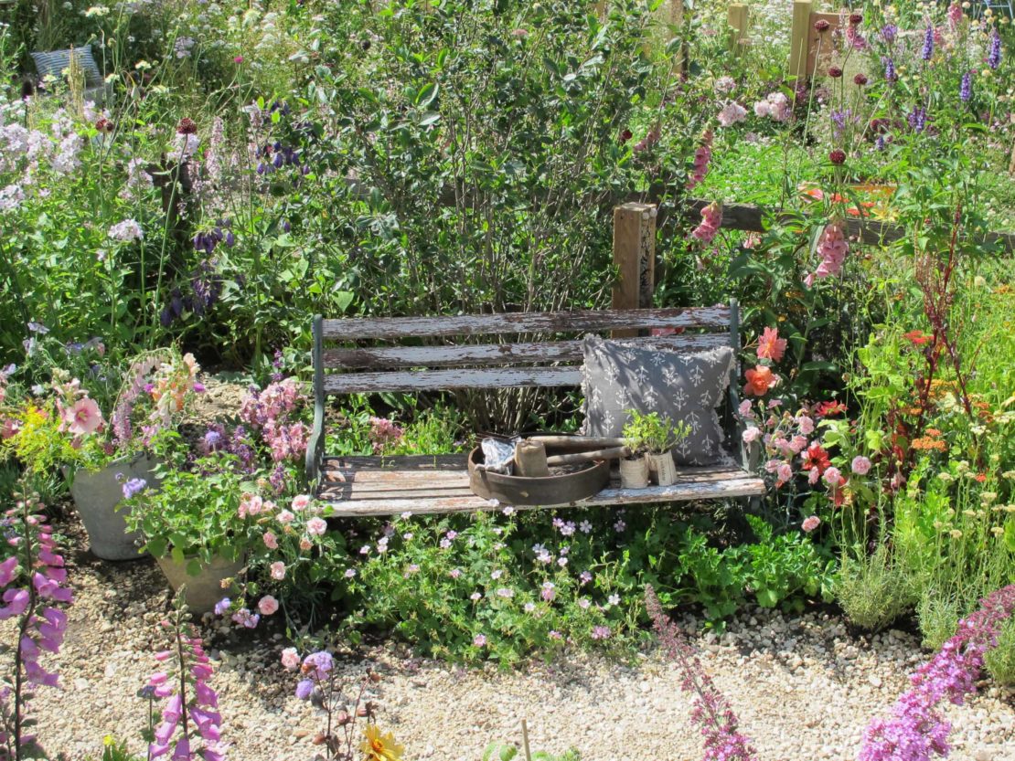 Eine Gartenbank, von blühenden Pflanzen umgeben, dahinter der Nachbargarten, der weder durch eine Mauer oder Hecke abgetrennt ist - solche lockeren Grenzen machen es Igeln leicht, von einem Garten zum anderen zu gelangen.