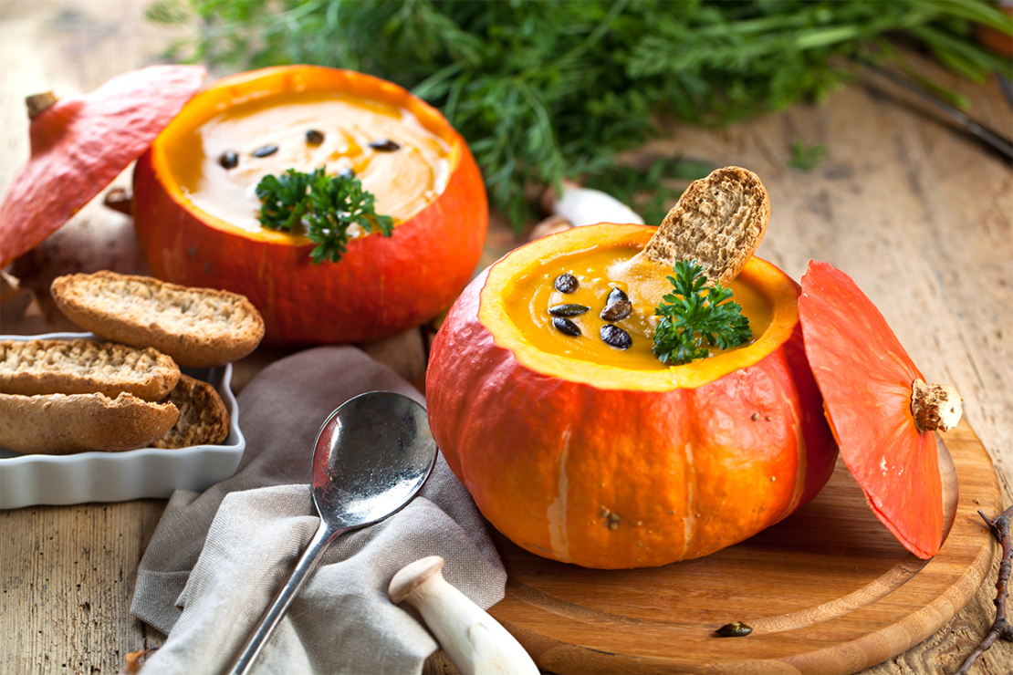 Kürbissuppe mit Orangensaft in eigener Schale auf einem Tisch mit Brot, Löffel und Kürbiskernen [Foto: AdobeStock_Karepa]