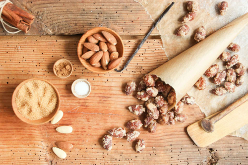 Gebrannte Mandeln in einer Tuete auf einem Holztisch neben Zucker und rohen Mandelkernen [Foto: AdobeStock_eflstudioart]