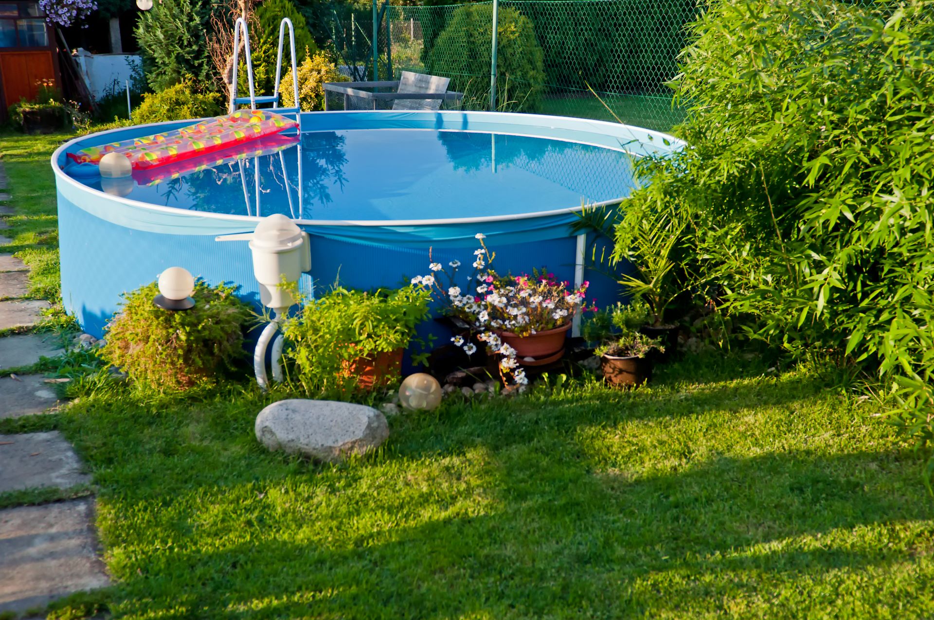 Mit Wasser gefüllter, runder Aufstellpool auf der Wiese eines Gartens. Pinkfarbene Luftmatratze schwimmt auf dem Wasser. Blumenkübel dekorieren den Poolbereich. 