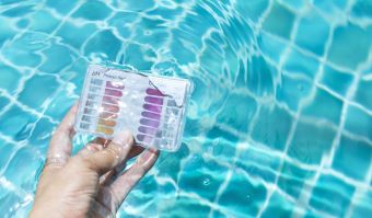 Eine Hand hält ein Test-Kit zur Überprüfung der Wasserqualität in das Wasser eines Pools.