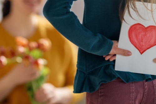 Mädchen versteckt eine selbstgemachte Karte mit einem roten Herz hinter ihrem Rücken, im Hintergrund eine Frau im beigen Oberteil, die einen Strauß Blumen in den Händen hält
