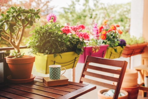 Gemütlich gestalteter Balkon mit Holzmöbeln im Vordergrund, auf dem Tisch ein geschlossenes Buch mit einer Tasse gemusterten Tasse darauf. Im Hintergrund bunte Balkonkästen mit bunt blühenden Stauden.