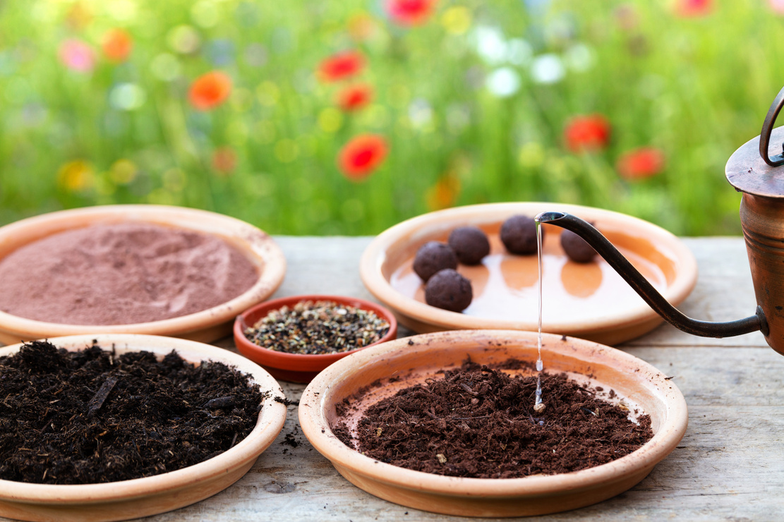 Tonteller gefüllt mit Blumenerde, Tonerde, Pflanzensamen und fertigen Samenbomben auf einem Holztisch vor blühender Wiese. Foto: AdobeStock_M.Dörr & M.Frommherz