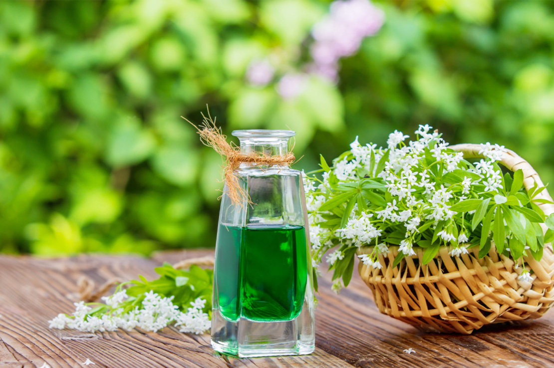 Waldmeistersirup in kleinem Glasfläschchen auf einem Holztisch. Rechts daneben steht ein kleiner geflochtener Korb mit gepflücktem Waldmeister. Foto: AdobeStock_fotoknips