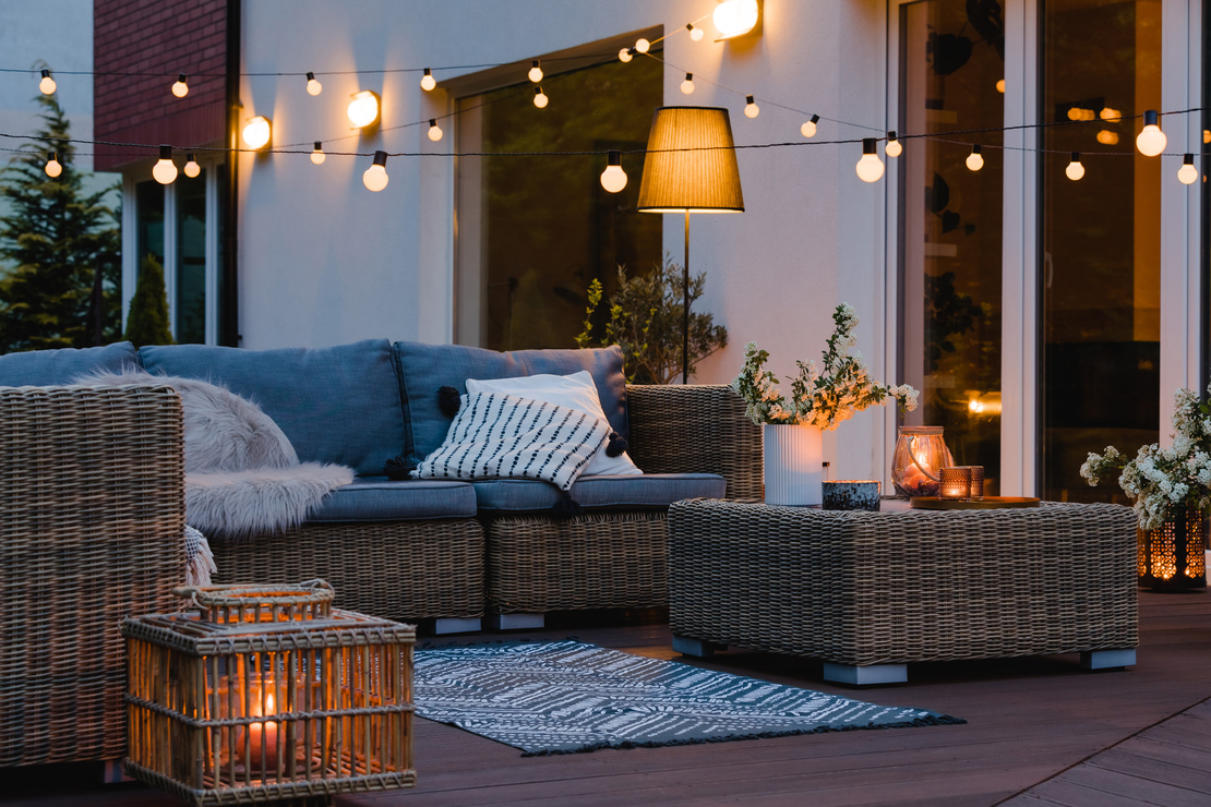 Smart-Gardening-Außenbeleuchtung. Eine Terrasse bei Abendstimmung: Korb-Gartenmöbel, eine Lichterkette, eine Stehlampe und eine Wandlampe. Foto: AdobeStock_Photographee.eu