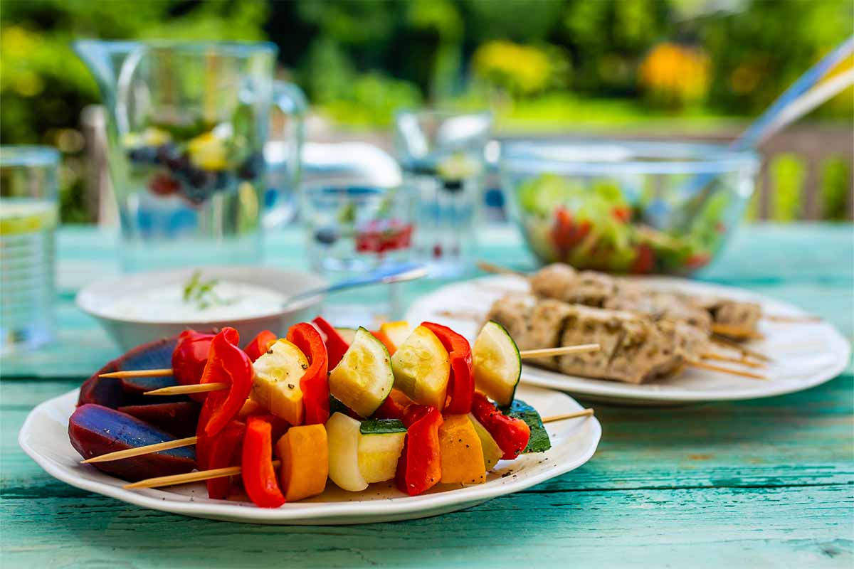 Grillspieße mit Gemüse und Fleisch neben einem gedeckten Gartentisch [Foto: AdobeStock_iMarzi]