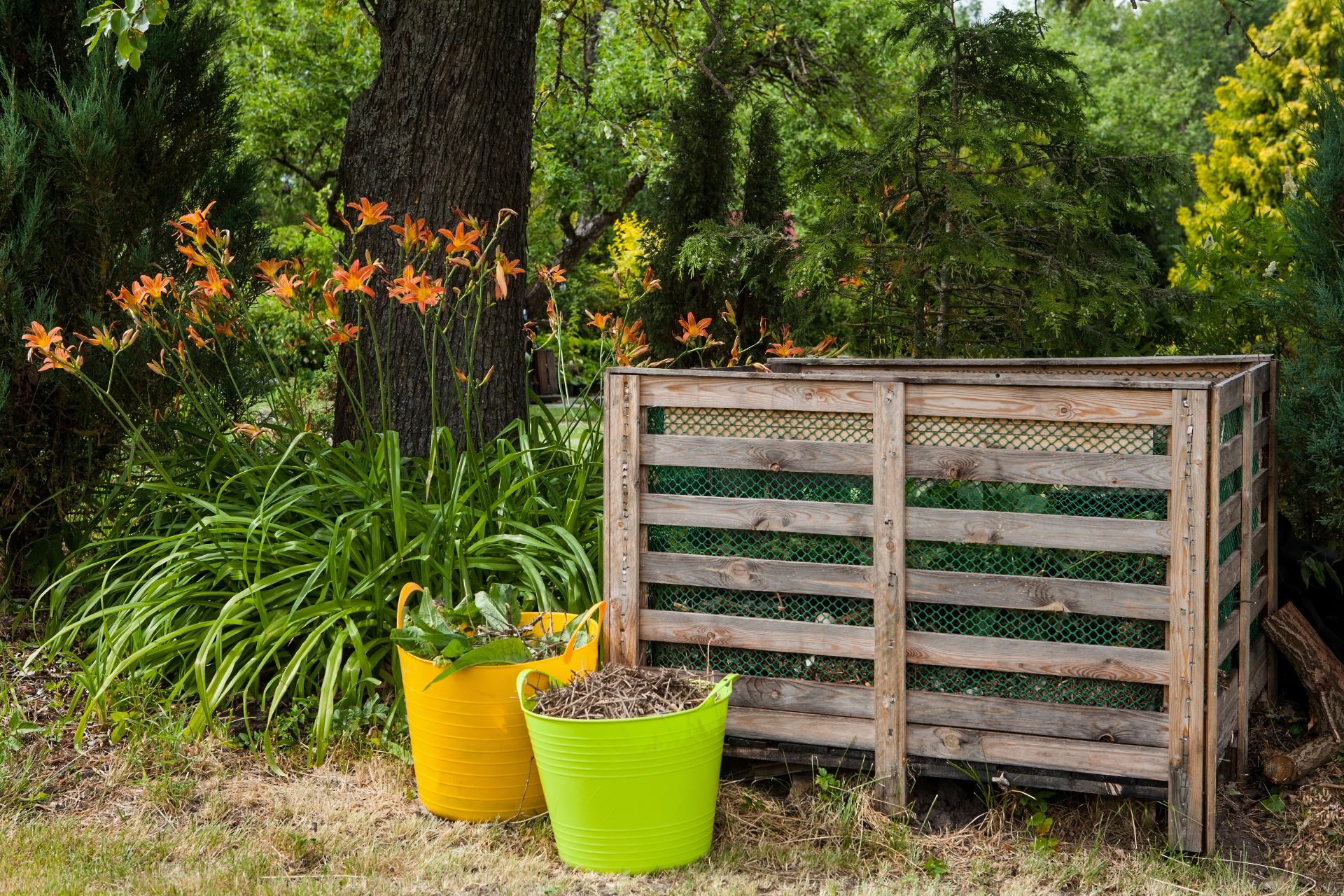 Komposter aus Holz, davor zwei Plastikbehälter in Grün und Gelb mit Gartenabfällen, stehen neben einem mit Lilien unterpflanzen Baum in einem Garten.