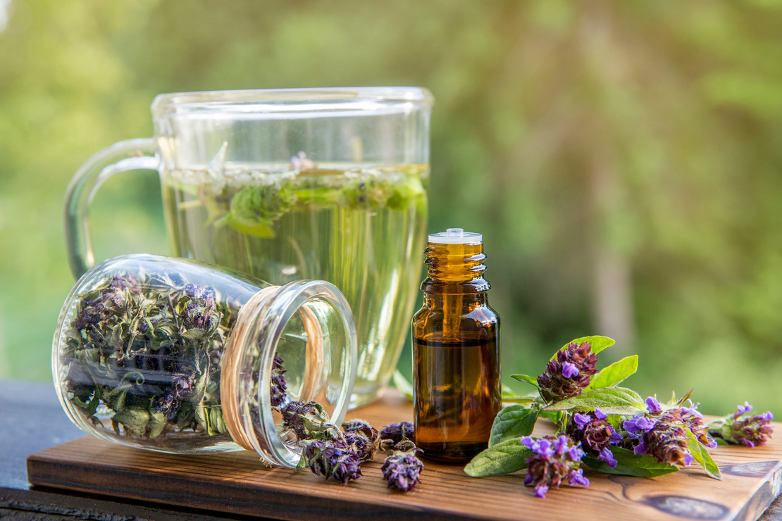 Aufgebrühter Tee mit Blättern der Kleinen Braunelle. Daneben ein kleines Fläschchen aus braunem Glas und ein Einmachglas mit Blüten der Kleinen Braunelle. Foto: AdobeStock_FotoHelin