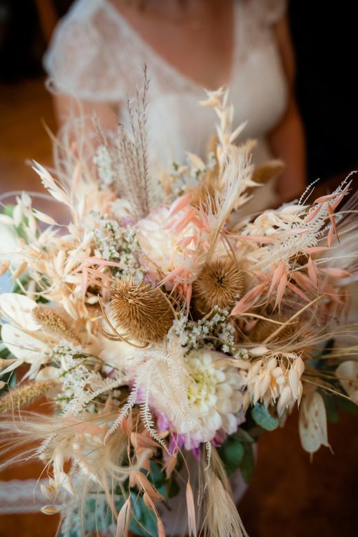 Brautstrauß aus Trockenblumen in zarten, pastellfarbenen Tönen. Im Hintergrund eine Frau im weißen Brautkleid, die den Blumenstrauß in der Hand hält.