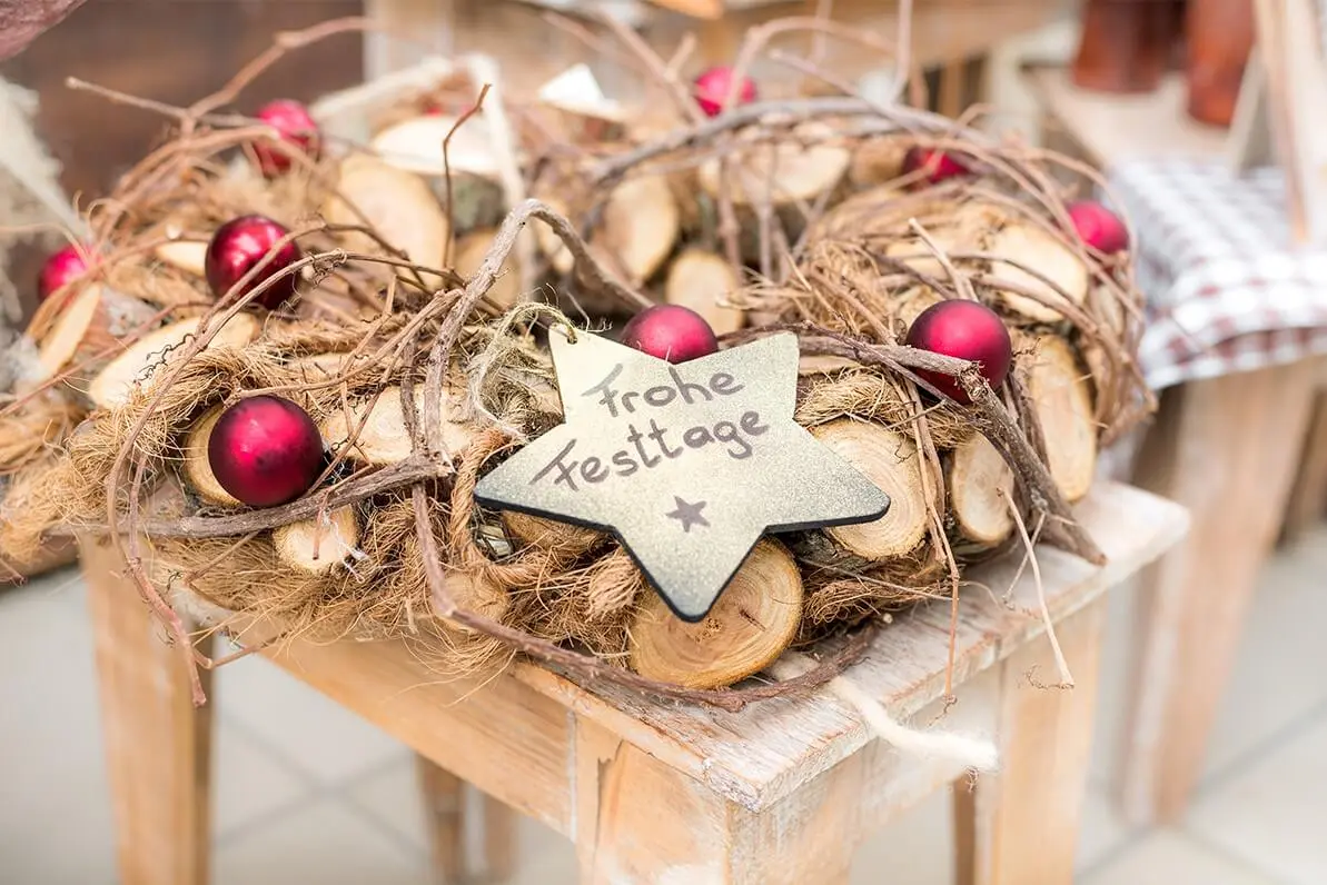 Winterdeko für den Garten mit einem Kranz aus Holz, der mit roten Weihnachtskugeln und einem Holzstern mit Feiertagsgruß verziert ist [Foto: AdobeStock_Lumixera]
