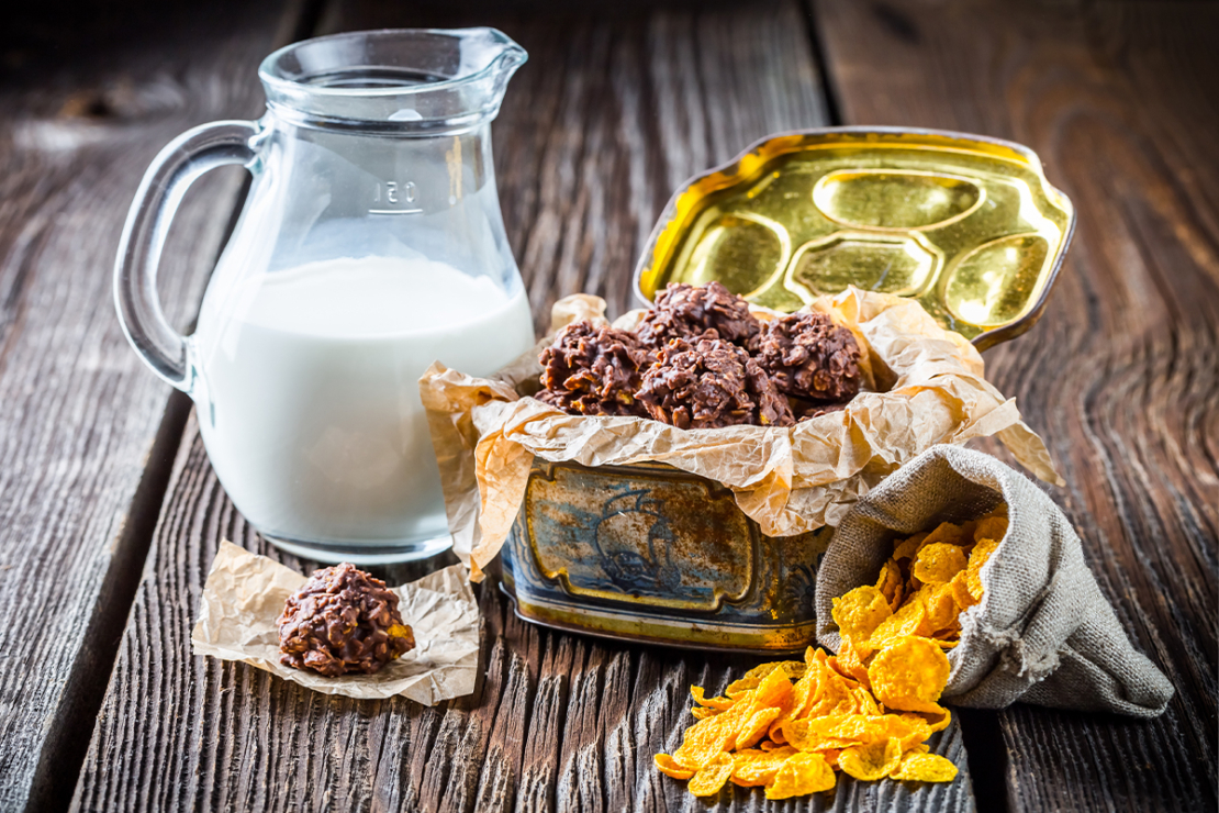 Schokocrossies in einer Blechdose neben Milch und einem Säckchen Cornflakes [Foto: AdobeStock_Shaiith]