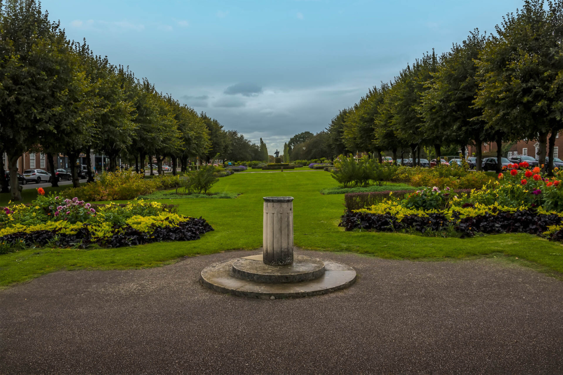 Gartenstadt: Blick inmitten eine Baumallee, zu beiden Seiten sind Blumenbeet, zentral im Bild eine Steinsäule. Foto: AdobeStock_Nicola