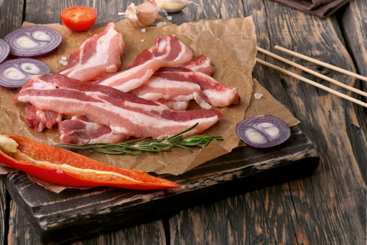 Grillfackeln werden aus Frühstücksspeck zubereitet – hier liegt Bacon auf einem Bettchen neben Spießen [Foto: AdobeStock_Kucherav]