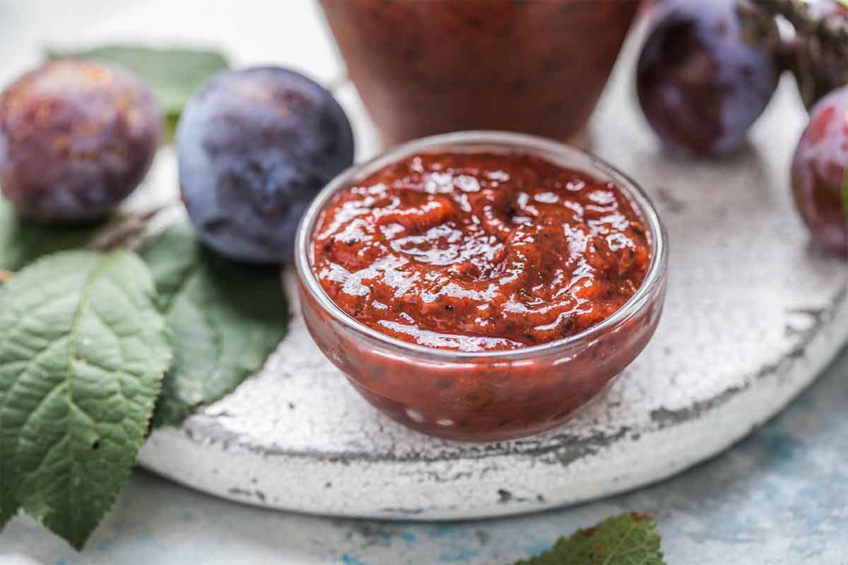 Glasschälchen mit Pflaumen-Chili-Sauce auf einem Untersatz aus Steingut. Im Hintergrund sind Zwetschken, Blätter und ein Krug zu sehen.  [Foto: AdobeStock_Maria]