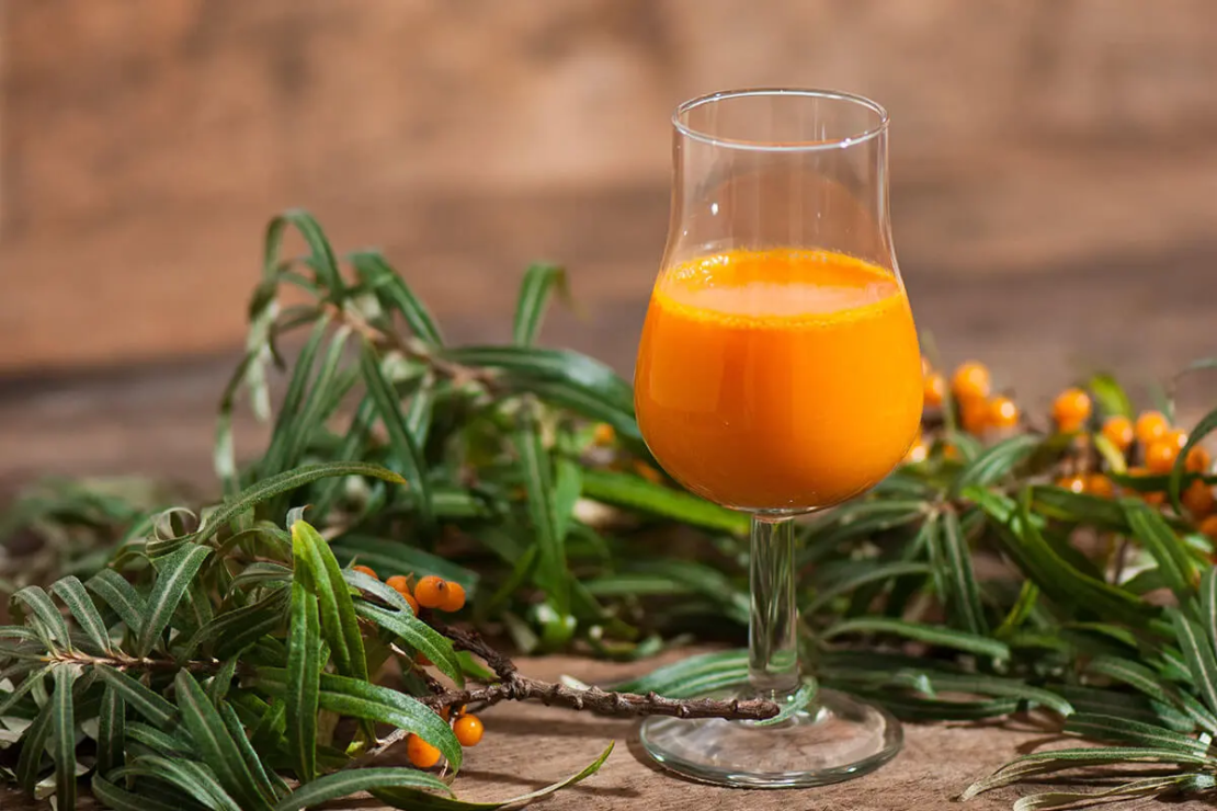 Sanddornsaft in einem Glas auf einem Tisch neben einem Sanddorn-Ast [Foto: AdobeStock_Superhasi]