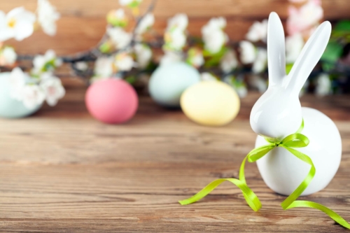 Eine Hasenfigur aus weißem Porzellan mit einer grünen Schleife um den Hals steht auf einem Holztisch, im Hintergrund bunte Ostereier und Blüten
