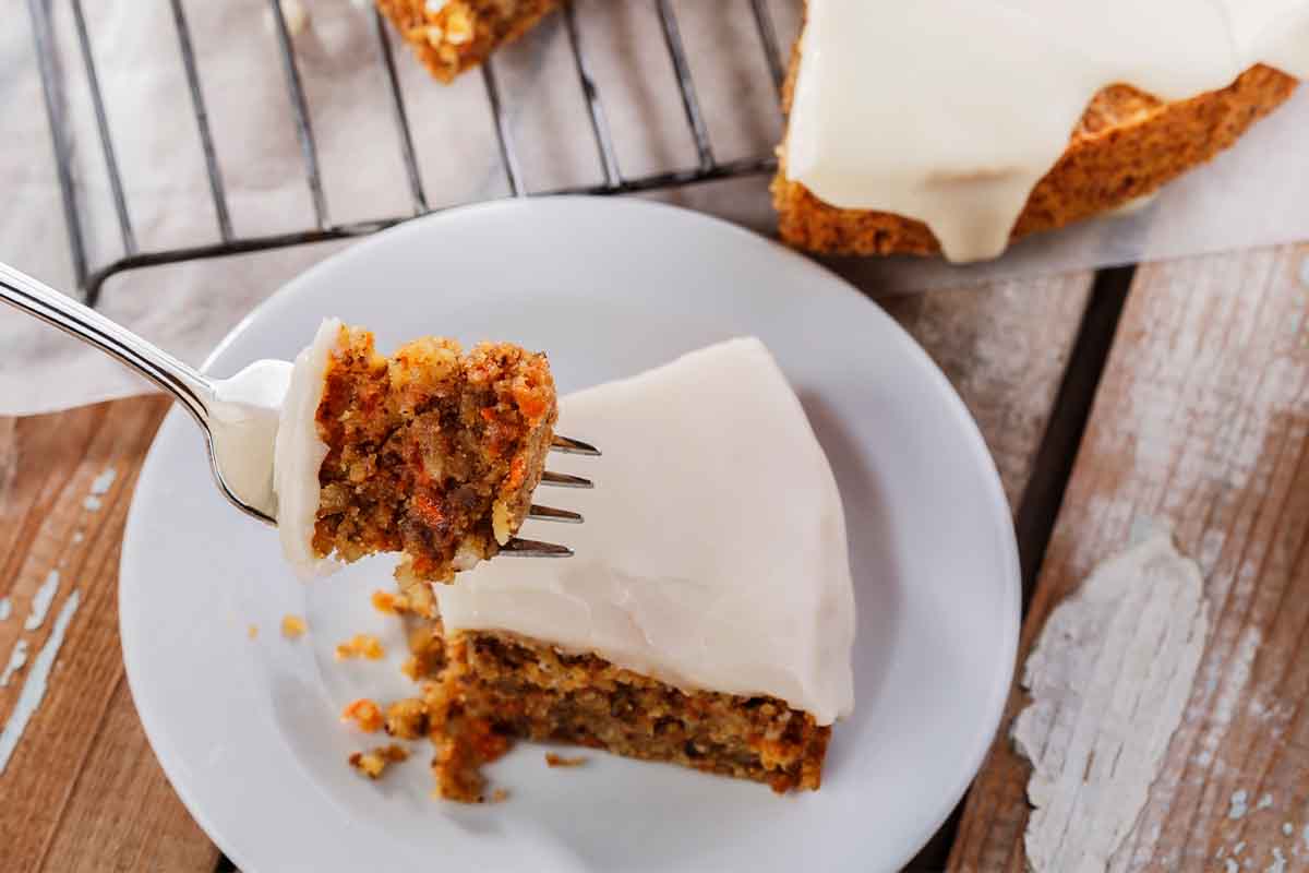 Rueblikuchen, der auf einem Kuchengitter in der Vogelperspektive fotografiert. Ein Stueck fehlt bereits und liegt auf einem weissen Teller in der unteren Bildhaelfte. Die vordere Spitze wurde aufgegabelt und ins Bild gehalten. [Foto: AdobeStock_koss13]