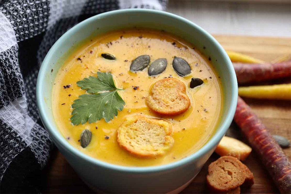 Suesskartoffelsuppe steht auf einem Tisch neben Karotten und Suesskartoffel und wurde in einem tiefen Teller angerichtet und mit Kuerbiskernen bestreut. [Foto: AdobeStock_Adrian]