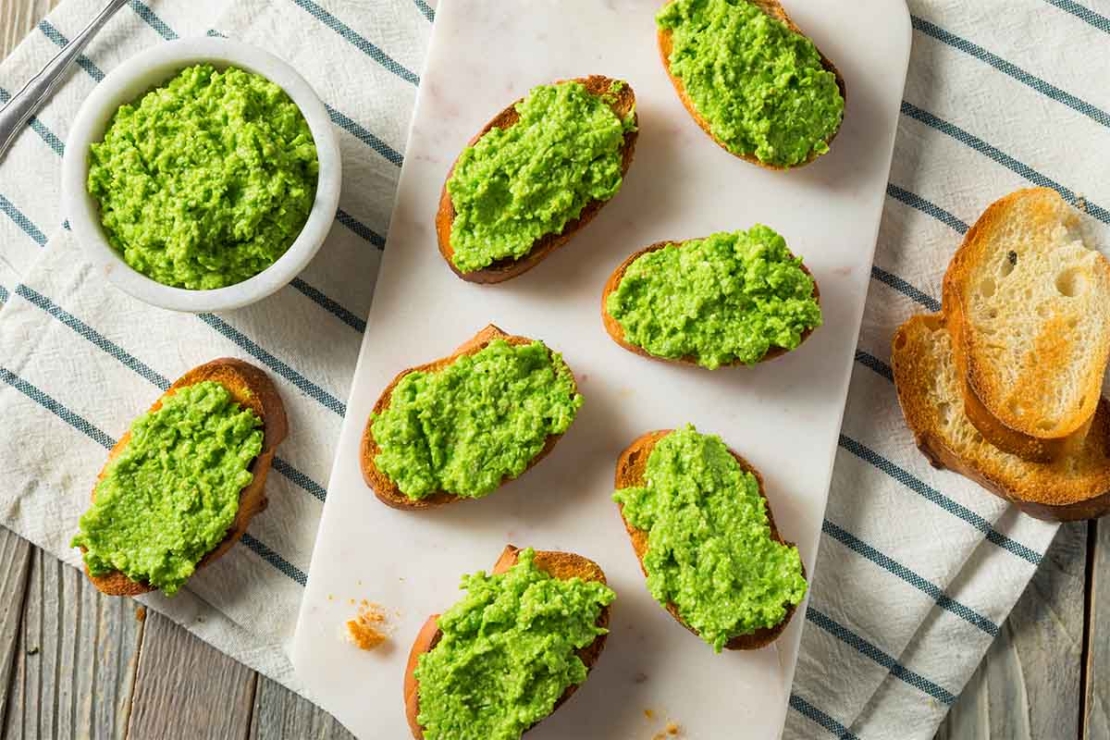 Erbsen-Pesto-aufstrich auf Baguette-Scheiben, die aus der Vogelperspektive fotografiert und nebeneinander auf einem Bettchen platziert wurden. Daneben steht ein Schaelchen mit dem Aufstrich, der herrlich gruen leuchtet. Die Tischdecke auf dem Tisch ist gruen-weiss gestreift. [Foto: AdobeStock_Brent Hofacker]