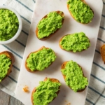Erbsen-Pesto-aufstrich auf Baguette-Scheiben, die aus der Vogelperspektive fotografiert und nebeneinander auf einem Bettchen platziert wurden. Daneben steht ein Schaelchen mit dem Aufstrich, der herrlich gruen leuchtet. Die Tischdecke auf dem Tisch ist gruen-weiss gestreift. [Foto: AdobeStock_Brent Hofacker]