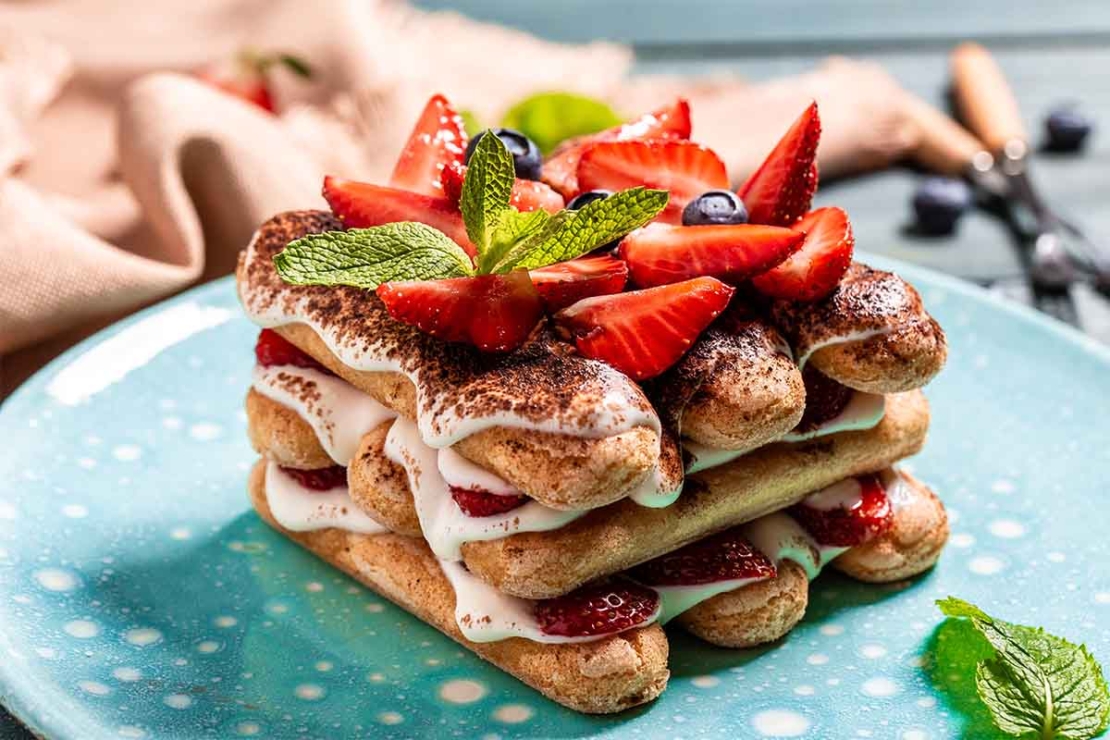 Erdbeer-Tiramisu, das mit Kakao bestäubt wurde und auf einem hellblauen Teller mit weissen Punkten serviert wird. [Foto: AdobeStock_Надія Коваль]