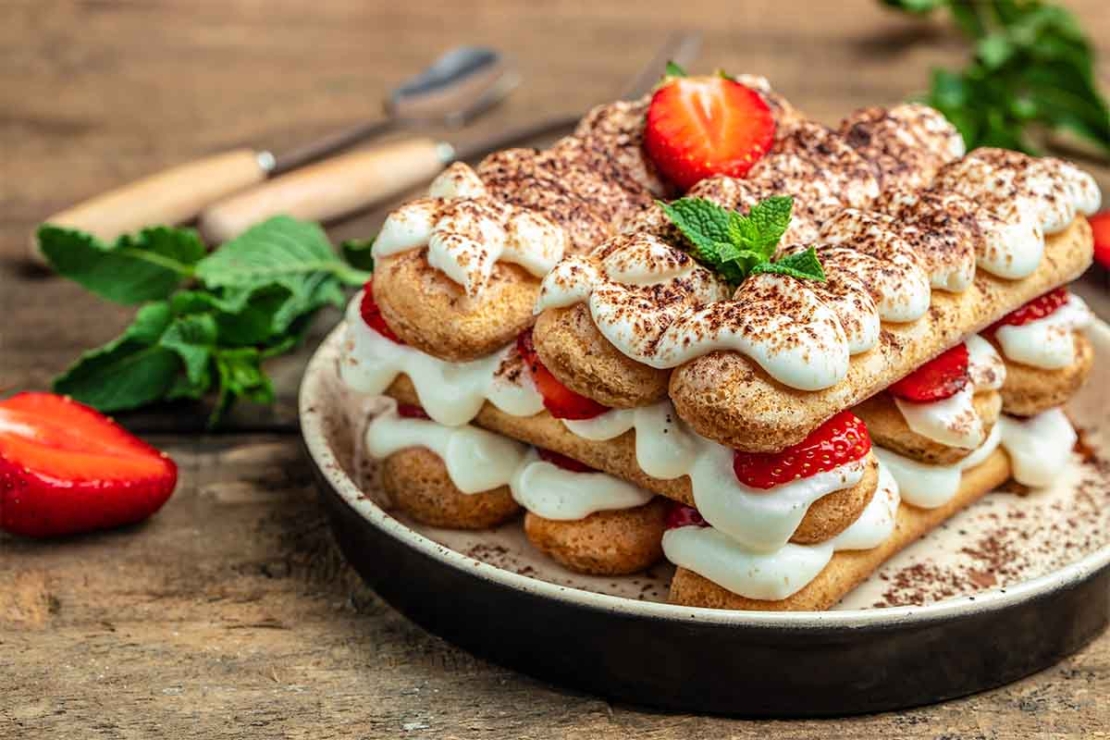 Zu sehen ist ein Holztisch, auf dem ein weisser Teller steht, auf dem eine Portion Erdbeer-Tiramisu angerichtet ist. Daneben liegt Besteck mit Holzgriffen. [Foto: AdobeStock_Надія Коваль]